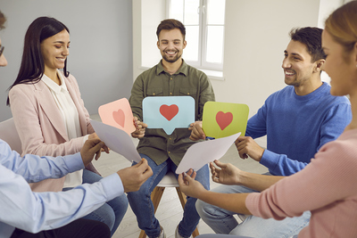 Group of happy people sitting in circle and holding paper message bubbles with heart symbols and like signs. Concept of young men and women communicating on social media, dating online, and having fun