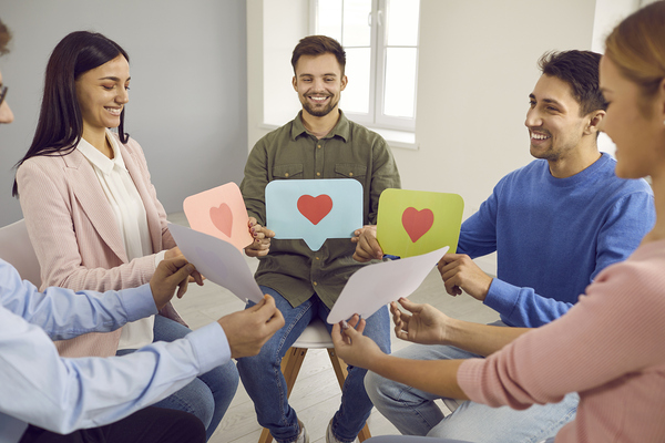 Group of happy people sitting in circle and holding paper message bubbles with heart symbols and like signs. Concept of young men and women communicating on social media, dating online, and having fun